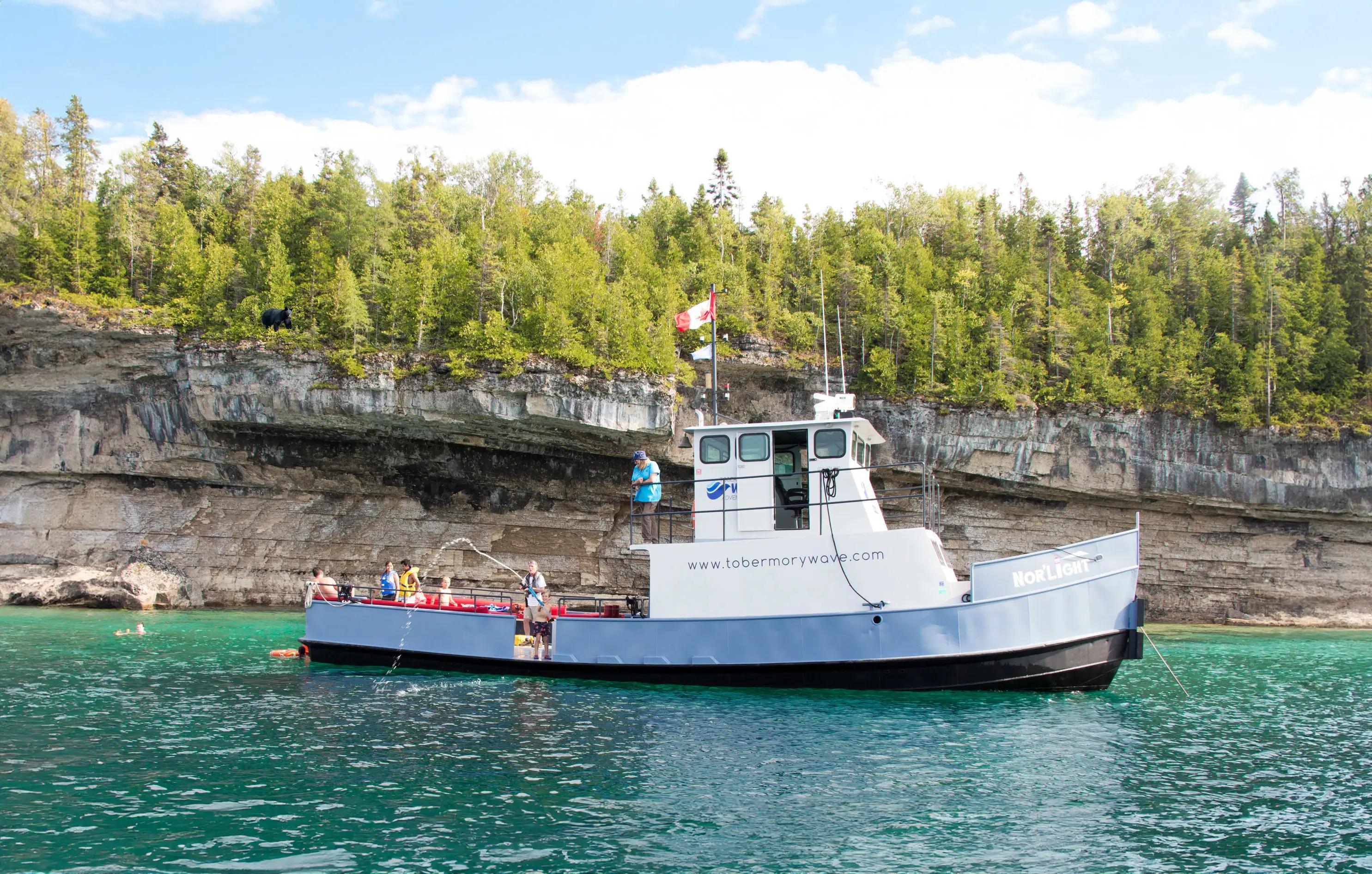 A classic-style tugboat anchored in front of a cliff, surrounded by clear water with people swimming around the boat.