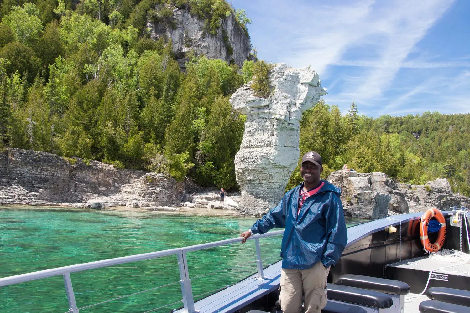 A man standing on an open-air tour boat that is in front or a rock formation known as a Flowerpot.