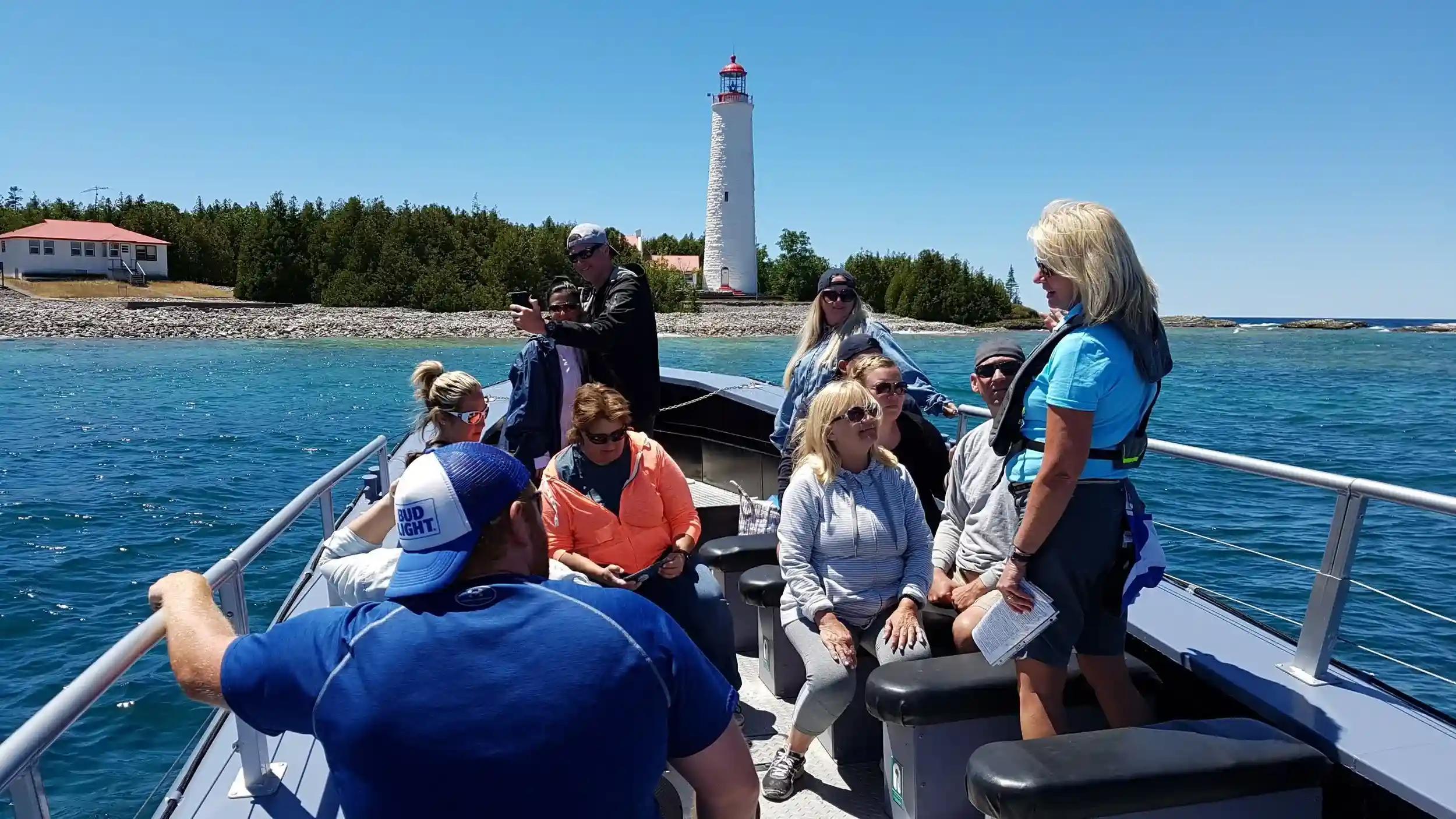 An open-air tour boat with a small group of people in front of a white and red brick lighthouse, known as Cove Island Lighthouse, on a clear, sunny summer day.